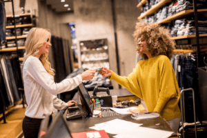 A smiling customer in a yellow jumper hands a credit card to a cashier behind the counter in a clothes shop filled with shelves of jeans.