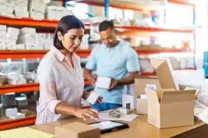 Two people work in a warehouse. A woman in front checks a tablet and handles a small box on a table with parcels, while a man in the background holds a box. Shelves with various boxes are visible behind them.