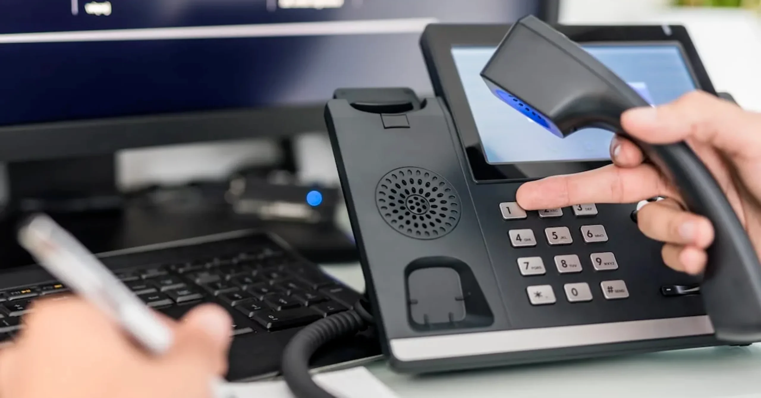 A person dials a number on an office desk phone while holding the handset, with a keyboard and computer monitor in the background. Another hand holds a pen, suggesting multitasking or note-taking.