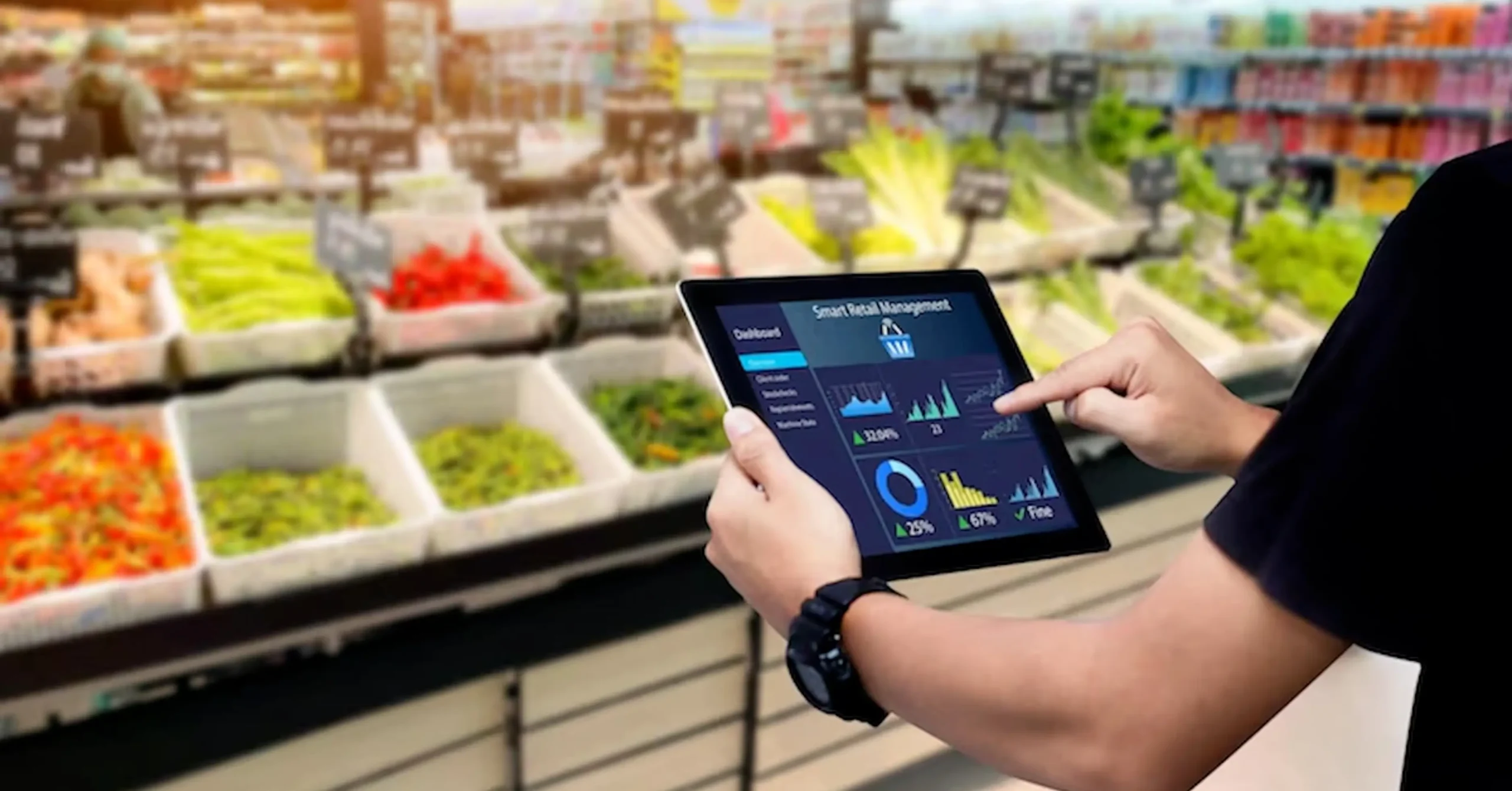 A person uses a tablet displaying data charts and analytics in a supermarket produce section with assorted fruits and vegetables in the background.