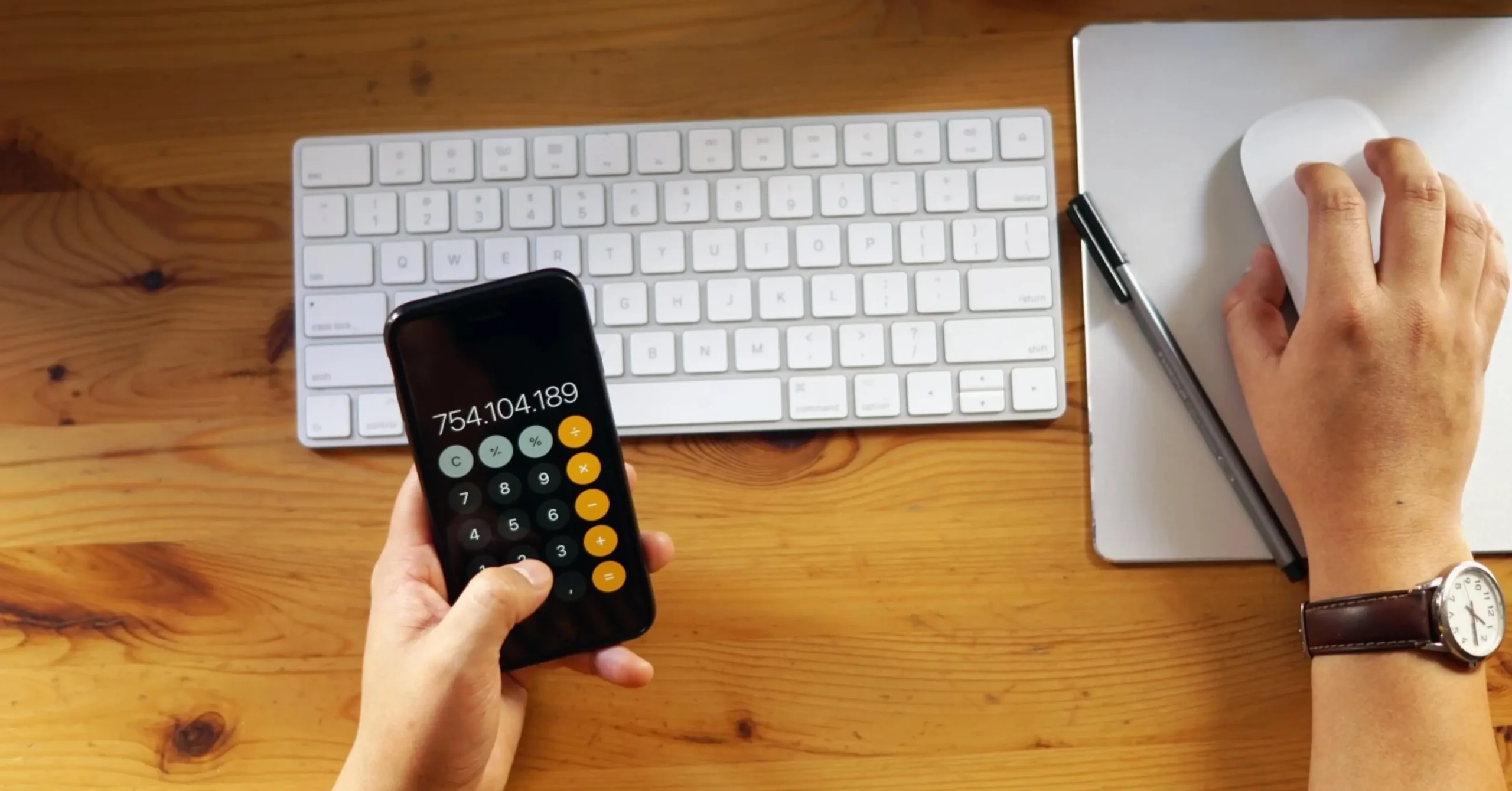 A person holding a smartphone with a calculator app open next to a keyboard, notebook, and pen on a wooden desk.