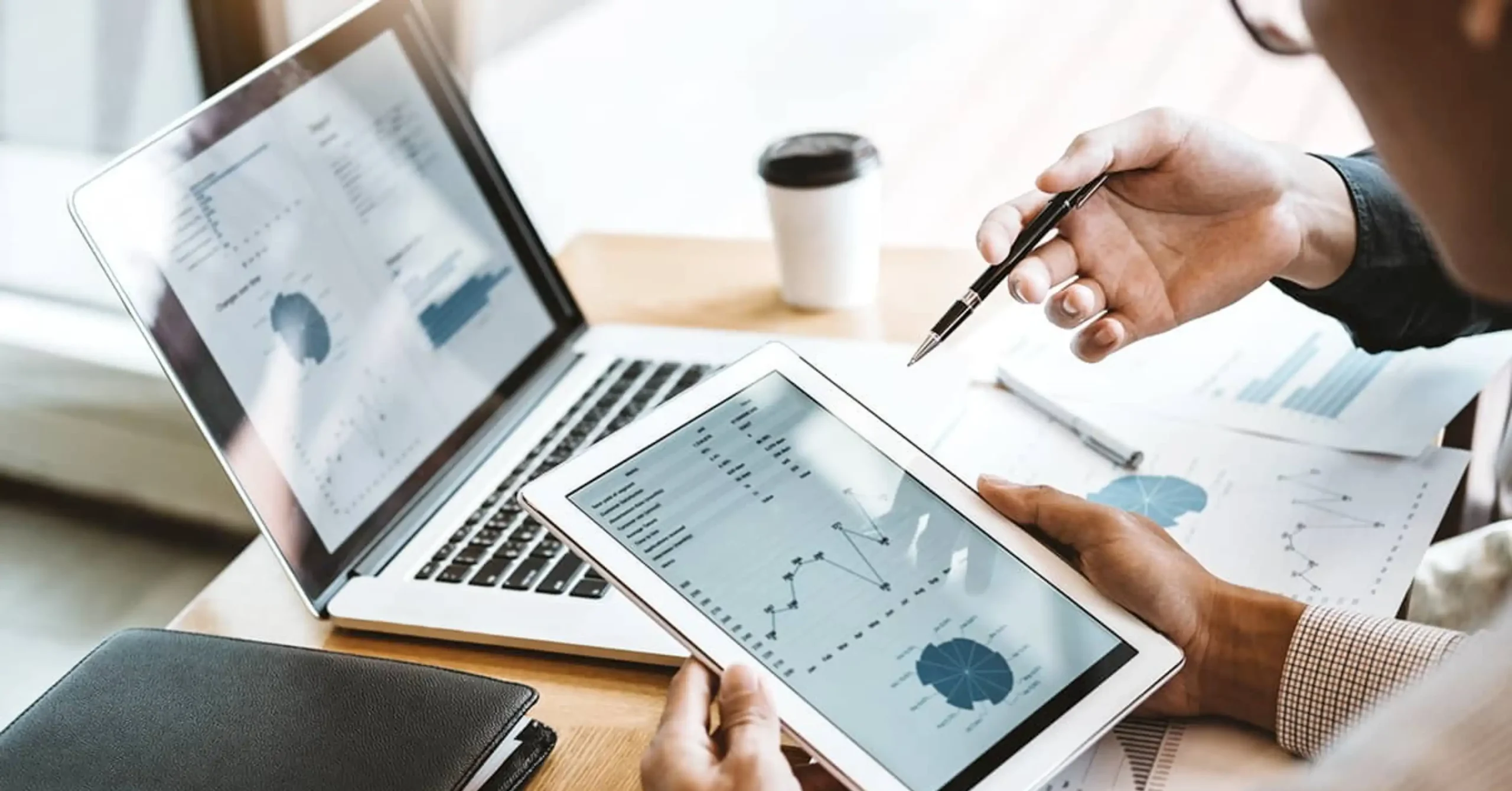 Two people review financial charts and graphs displayed on a tablet and a laptop at a desk, with one person pointing at the tablet. A coffee cup and documents are also on the desk.